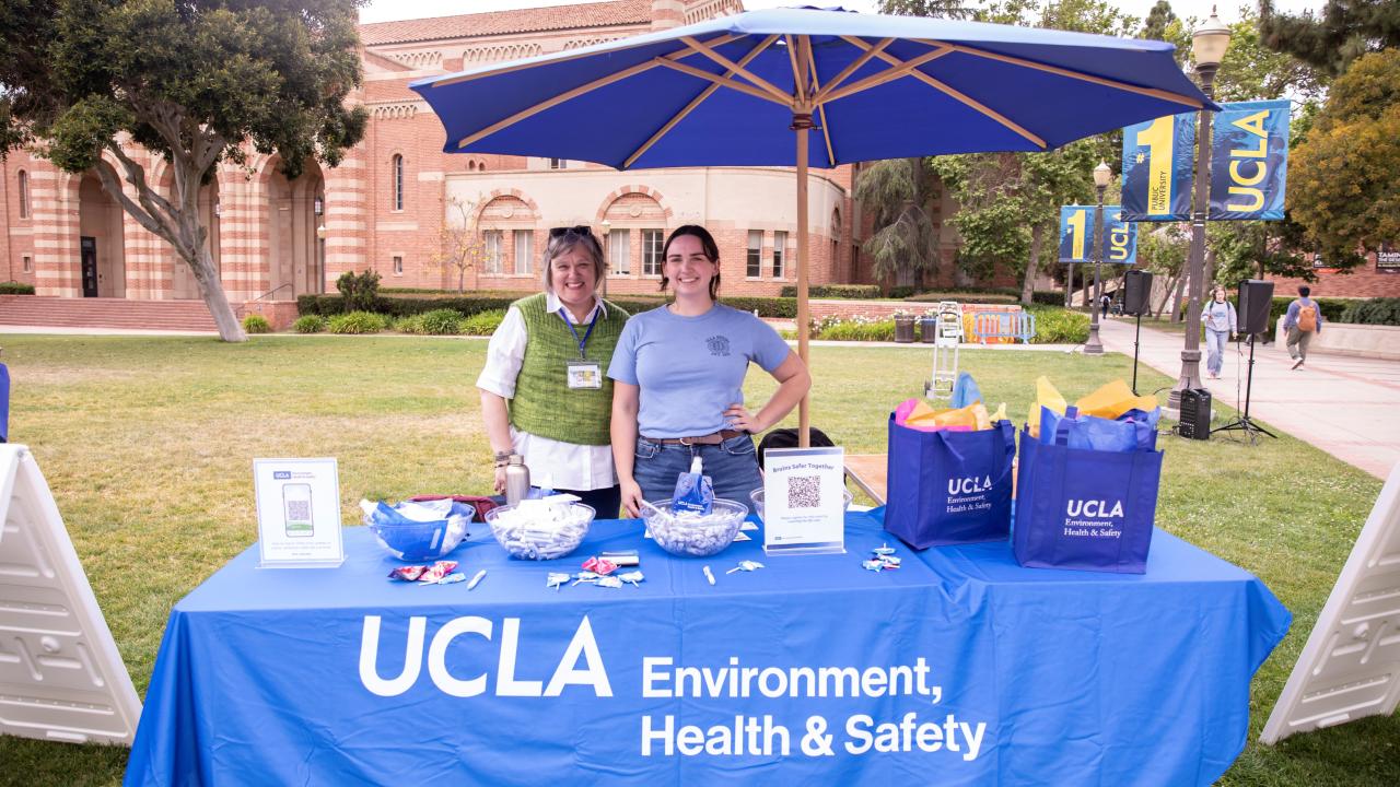 Two women behind a table with the UCLA Environment Health & Safety Logo and gift bags on top