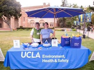 Two women behind a table with the UCLA Environment Health & Safety Logo and gift bags on top